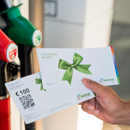 Hand hält Kostner Tankgutschein über 100 Euro mit QR-Code vor der Zapfsäule – perfektes Geschenk für Südtirol-Autofahrer